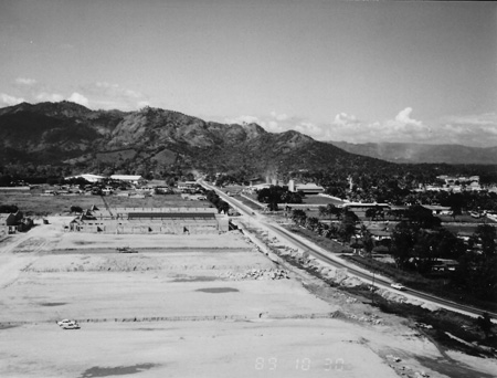 Aerial black-and-white photograph of a cleared site and buildings under construction beside a highway, with mountains in the background